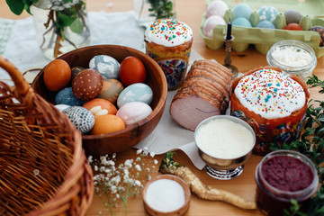 Obraz na płótnie Canvas Easter preparations. Easter modern eggs, easter bread, ham, beets, butter, green branches and flowers on rustic wooden table with wicker basket and candle. Traditional Easter Food