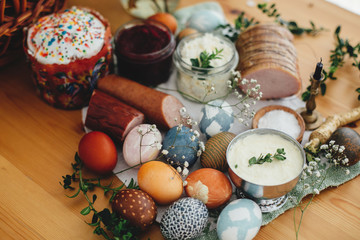 Fototapeta na wymiar Easter modern eggs, easter cake, ham, beets, butter, cheese, sausages, green branches and flowers on rustic wooden table. Holiday preparation. Traditional Easter Food for breakfast