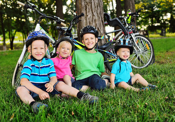 Fototapeta premium a group of small preschool children in Bicycle safety helmets smile sitting on the fresh green grass in the Park against the background of bicycles and trees.