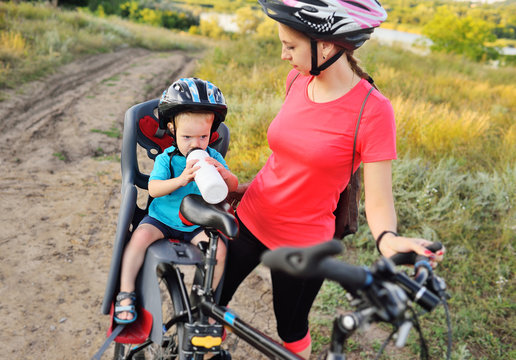 a small child a boy in a protective helmet sitting in a child's Bicycle seat drinking water from a bottle on the background of a large Bicycle and his mother. Bike ride.