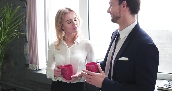 Beautiful Blond Woman Trying To Seduce Youn Coworker, Flirting With Him While They Drink Coffee Near Window At Workplace