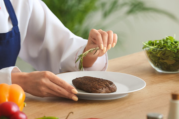 Female chef adding herbs to tasty meat in kitchen, closeup