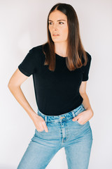 Indoor portrait of beautiful young woman, wearing black t-shirt and high waist jeans, posing on white background, holding hands in pockets