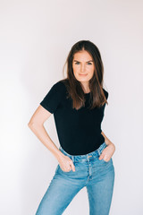 Indoor portrait of beautiful young woman, wearing black t-shirt and high waist jeans, posing on white background, holding hands in pockets