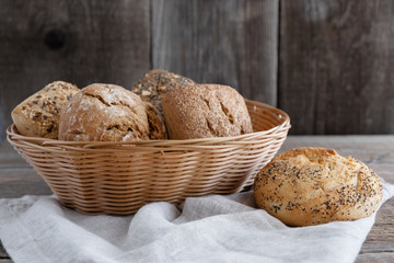Different varieties of bread and bun lie in a wicker basket and next to them are a wheat roll and a linen table napkin. Rye, spelled, whole grain baked goods with sunflower and chia seeds.