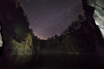 The stars above an abandoned tree lined quarry with partially lit cliffs.
