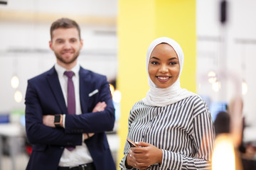 Fototapeta na wymiar African american muslim girl with hijab posing with Caucasian business colleague wearing suit in modern office.
