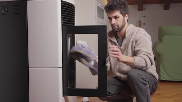 Young man cleaning new pellet stove door glass inside warm apartment 's living room interior with wooden floor. Eco ideas for house heating. Wood chip / pellet stove maintenance and cleaning. 1 of 2 