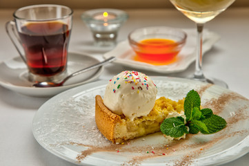 Piece of cake charlotte with apples ice cream ball and fresh mint on white plate. Served with a rosette of honey, a cup of aromatic tea and a glass of white wine