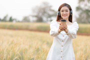 Fototapeta premium Young beautiful woman with coffee drink standing outdoors in barley field land and listening the music by wireless headsets in the morning.