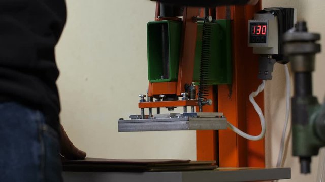 Close-up of craftsman's hands using press machine to stamp logo on pattern of leather product during work in crafting atelier. Male worker manufacturing exclusive leather goods in craft workshop