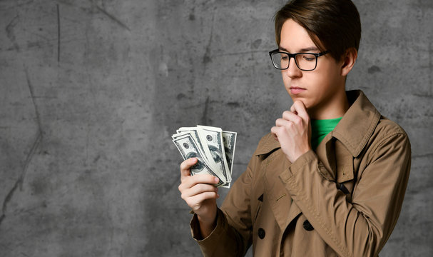 Young smiling teen boy in brown jacket and glasses standing, looking at heap of dollars in hand and thinking what to do