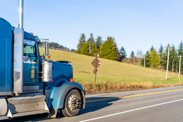 Blue big rig classic semi truck with chrome and aluminum accessories running on the road with meadow on the hill