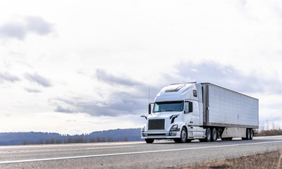 Industrial transportation big rig white semi truck transporting frozen cargo in reefer semi trailer running on the road with cloudy sky