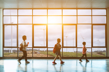 A group of children walking in the building There is a lot of large wind  turbine outside the window.