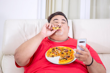 Fat man using his smartphone while biting a pizza