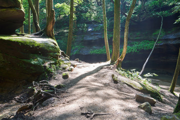 A trail in Hocking Hills State Park traveling near the river through a rock lined valley, with rock cliffs on the side.