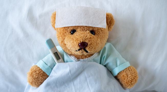 The teddy bear is sick on the bed with a high fever. There is a fever reducing sheet on the forehead.
