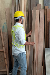 Handsome man wearing safety vest and put yellow helmet on head,choosing timber board,doing wood work at factory,Handmade design