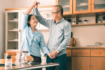 Asian  senior couple is dancing and smiling while cooking together in kitchen.