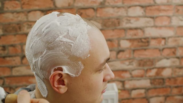 Barber applying foam with brush on skin bald man before shaving in male salon. Male stylist preparing client to shaving head with straight razor in barbershop. Skin and hair care