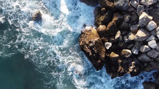 A birds eye top down view of powerful azur blue Mediterranean sea waves crashing and splashing against coastal rocks with lots of spray during a breezy sunny day in the south of France in Beausoleil.