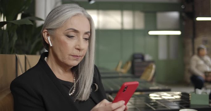 Close up of old female businessowner in earpods using smartphone and typing. Modern mature woman sitting in cafe while writing message and smiling. Concept of people, communication .