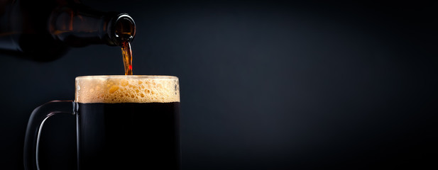A mug of black beer pouring from a bottle close-up, on a dark background. Stock photo.