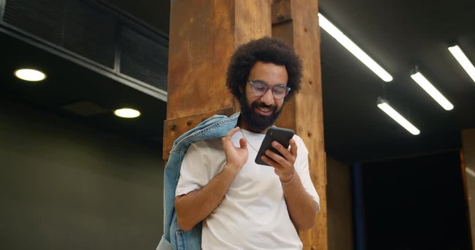 Bottom view of handsome guy in glasses laughing while using his smartphone. Bearded guy in casual clothes standing leaned to metal support and chatting in social networks.