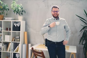 Young male architect standing in modern office with cup of coffee in his hand.	