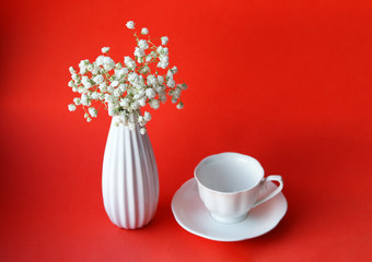A beautiful ceramic white vase with a bouquet of white gypsophila flowers and a tea Cup with a white saucer on a red background. Selective focus. Interior decorating.