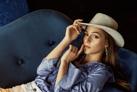 Fashion portrait of young elegant, luxury woman wearing stylish white hat, trendy pearl hoop earrings, blue silk blouse 