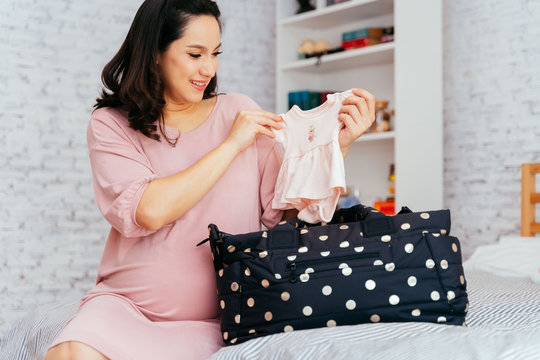 Asian Mixed Caucasian Pregnant Woman Packing Clothes Into A Bag For Expacting Baby To Be Born. Healthy And Happy Mother To Be Prepares A Newborn Shirt For Baby.