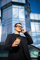 Young successful businessman wearing black suit and talking on his smartphone while standing near modern office or skyscrapers