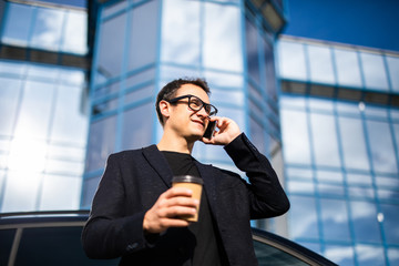 Handsome professional fresh stylish modern business man calling on cellphone with coffee near skyscraper