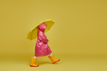 Cheerful child girl with yellow umbrella and red rain coat on colored yellow background. Copy space...