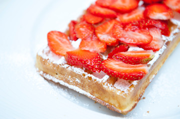 Fresh gourmet Belgian waffle served on a place with powdered sugar and fresh strawberry slices; selective focus on near corner 