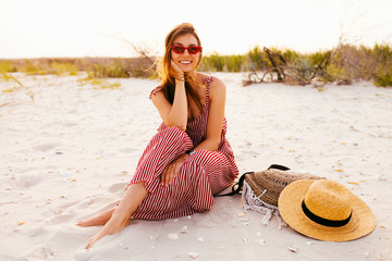 Horizontal  image of boho style young woman relaxing on the beach. Graceful young woman in trendy summer linen straw romper and straw hat posing on the beach near ocean. Summer style. Boho bag.