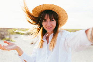 Close up of cheerful young caucasian  girl in summer hat taking a selfie at the beach, wearing white shirt and straw hat.Beautiful cheerful young woman having a good time at the beach on a lovely day,