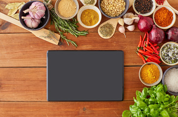 food, culinary and eating concept - tablet pc computer among different spices, onion, garlic with pine nuts and red hot chili peppers on wooden table