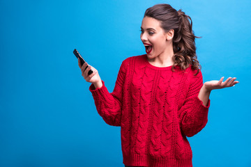 Emotional portrait of brunette caucasian pretty woman with evening make up and curly ponytail. She wears red sweater and poses on blue background with her phone in hand, having good news. Copy space