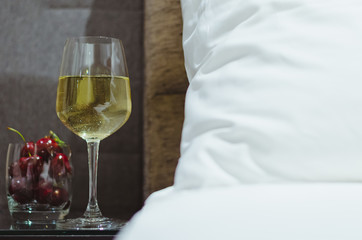 A glass of white wine with cherry fruits put on table besides bed with white bedsheet.