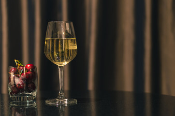 A glass of white wine with cherry fruits put on table in the room with curtain background.