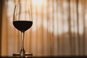 A glass of red wine on table in the room with sunset light on curtian background.