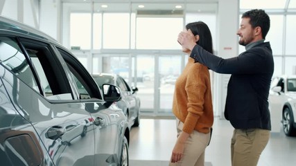 Businessman is buying new car for attractive young girlfriend making exciting surprise, happy lady is hugging boyfriend and laughing enjoying present.