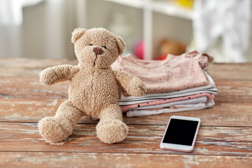 babyhood, technology and clothing concept - baby clothes, teddy bear toy and smartphone on wooden table at home