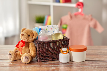 babyhood and care products concept - baby things in wicker basket and teddy bear toy on wooden...