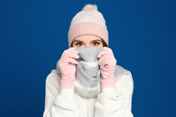 Young woman wearing warm sweater, gloves, scarf and hat on blue background. Winter season