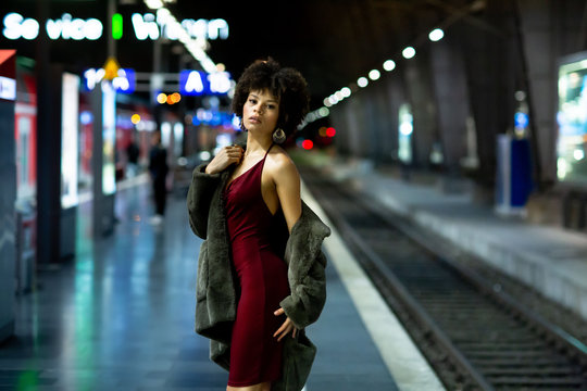 Young stylish African-American model woman in red dress and a fur coat posing on the subway platform at night. Fashion and lifestyle concept. Nightlife in the city