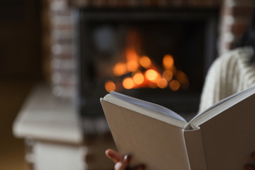 Woman reading book near burning fireplace at home, closeup